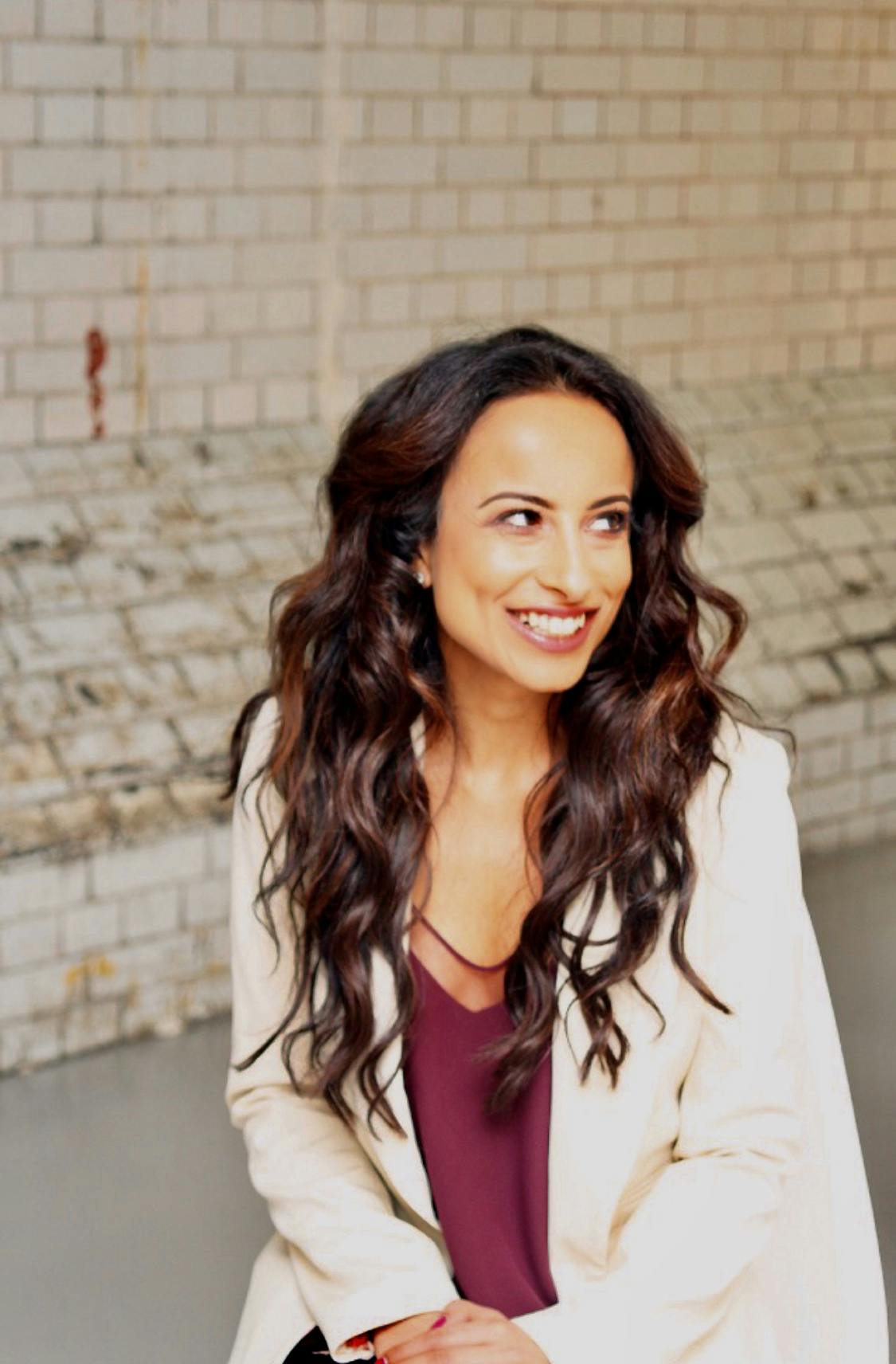 Woman, light-skinned with wavy hair. Wearing a white blazer with a burgundy undervest. Smiling.